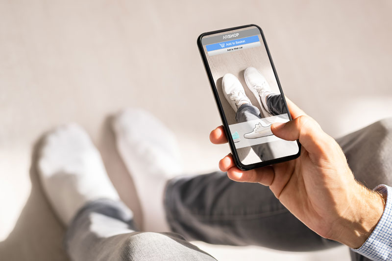 Person nutzt eine Augmented-Reality-App auf dem Smartphone, um weiße Sneaker virtuell an den eigenen Füßen auszuprobieren.